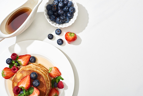 top view of delicious pancakes with blueberries and strawberries on plate near maple syrup in gravy boat on marble white surface