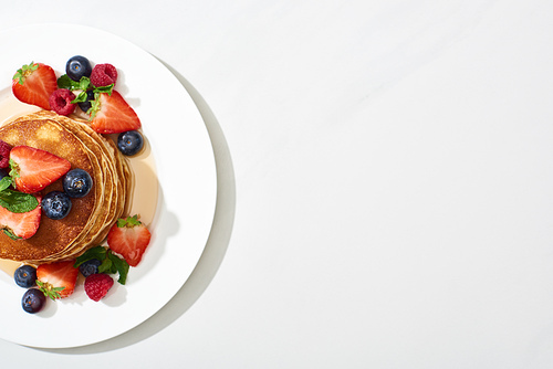 top view of delicious pancakes with maple syrup, blueberries and strawberries on plate on marble white surface