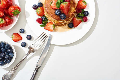 top view of delicious pancakes with honey, blueberries and strawberries on plate with cutlery on marble white surface