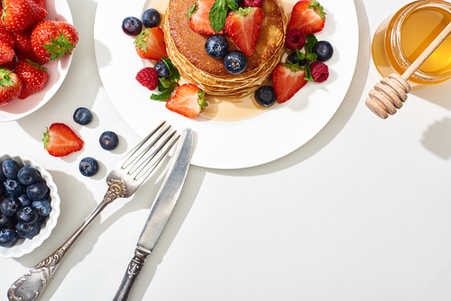 top view of delicious pancakes with honey, blueberries and strawberries on plate with cutlery on marble white surface