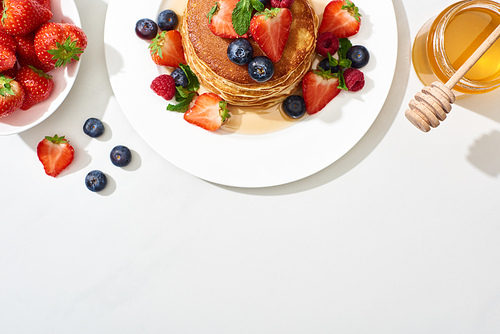 top view of delicious pancakes with honey, blueberries and strawberries on plate on marble white surface