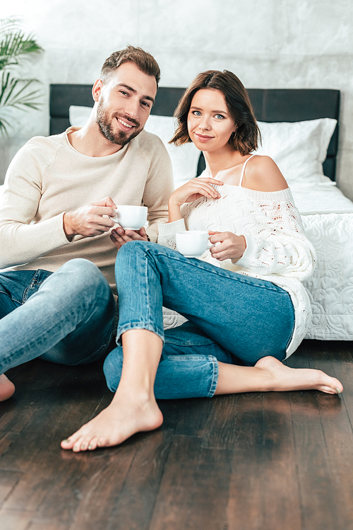 handsome man and beautiful woman sitting on floor and holding cups with drinks