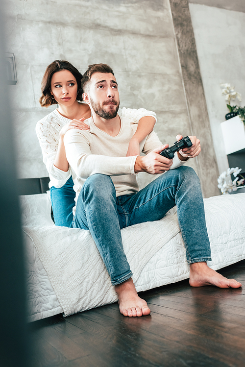 low angle view of brunette woman hugging handsome man sitting on bed and playing video game