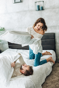 cheerful woman having pillow fight with happy bearded man on bed