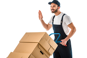 happy delivery man transported cardboard boxes loaded on hand truck and showing hello gesture isolated on white