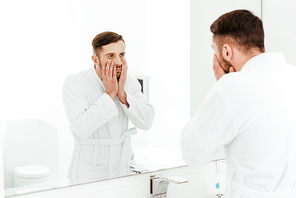 selective focus of upset bearded man looking at mirror while touching face in bathroom
