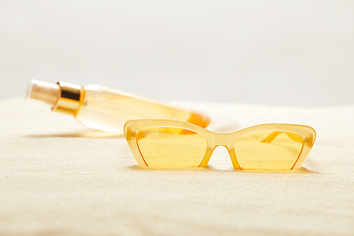 selective focus of yellow sunglasses near suntan oil bottle on sand isolated on grey