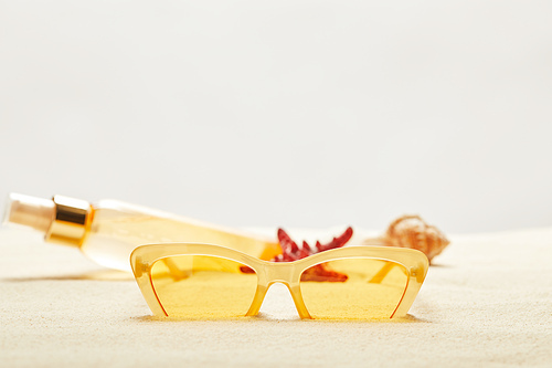 selective focus of yellow sunglasses near suntan oil bottle on golden sand isolated on grey