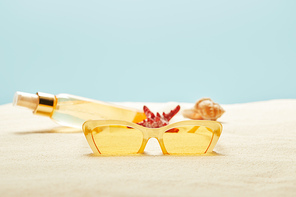 selective focus of sunglasses near suntan oil bottle, red starfish and seashell on sand  isolated on blue
