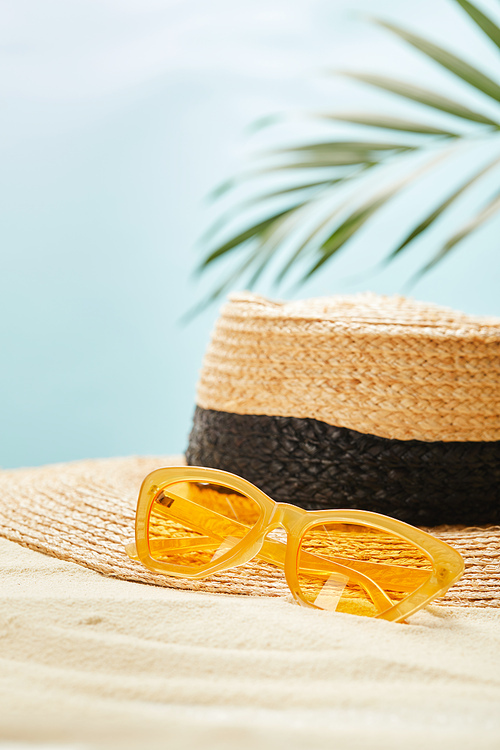 selective focus of yellow sunglasses near straw hat on golden sand in summertime isolated on blue