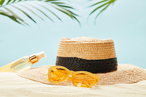 selective focus of yellow sunglasses near straw hat and bottle with suntan oil on sand isolated on blue