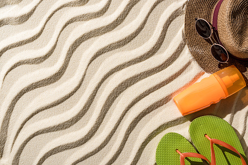 top view of straw hat, flip flops, sunscreen and sunglasses on wavy sand with copy space