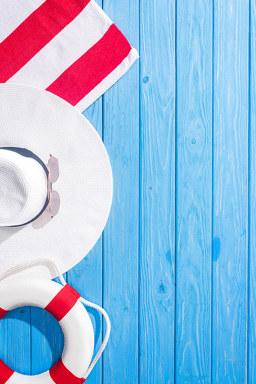 top view of striped towel, white sunglasses, lifebuoy, floppy hat on blue wooden background with copy space