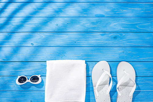 top view of white folded towel, retro sunglasses and flip flops on blue wooden background with shadows and copy space