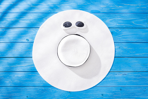 top view of white sunglasses and white floppy hat with black ribbon on blue wooden background