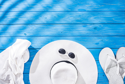 top view of crumpled towel, white glasses, flip flops and floppy hat on blue wooden background with copy space