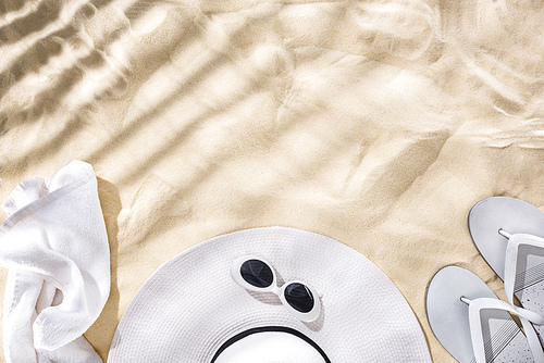 top view of white towel, retro sunglasses, flip flops and floppy hat with black ribbon on sand with shadows and copy space