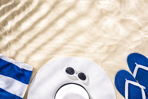 top view of striped towel, retro sunglasses, floppy hat and blue flip flops on sand with shadows and copy space