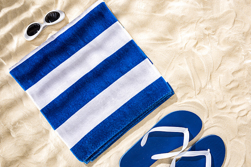 top view of white blue striped folded towel, retro sunglasses and flip flops on sand
