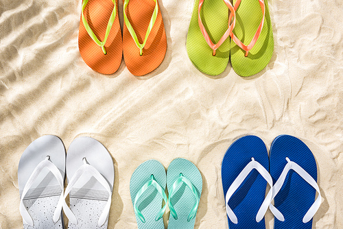top view of white, green, orange, turquoise and blue flip flops on sand with shadows