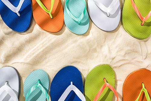 top view of white, green, orange, turquoise and blue flip flops on sand