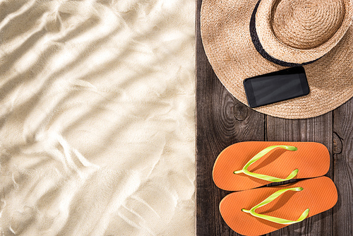 top view of sand with copy space and straw hat with black ribbon, smartphone and orange flip flops on wooden brown board