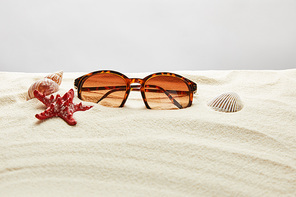 brown stylish sunglasses on sand with red starfish and seashells on grey background