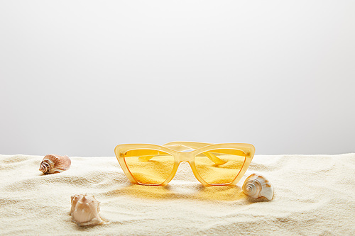 yellow stylish sunglasses on sand with seashells on grey background