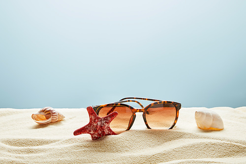 brown stylish sunglasses on sand with seashells and starfish on blue background