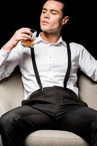 low angle view of handsome man holding glass of whiskey and sitting in armchair isolated on black