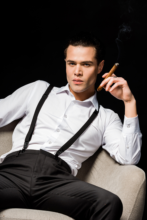 handsome man holding cigar while sitting in armchair isolated on black