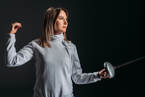 Beautiful swordswoman in fencing suit holding rapier and looking away isolated on black