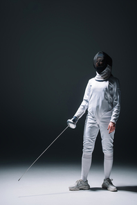 Fencer in fencing mask holding rapier while standing on white surface on black background