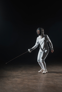 Swordswoman in fencing suit and mask training with rapier on black background