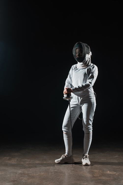 Fencer in fencing mask and suit holding rapier on black background