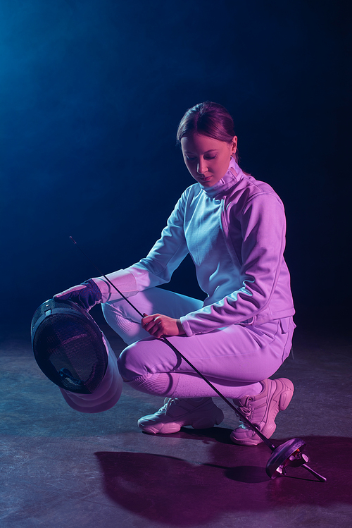 Beautiful fencer holding fencing mask and rapier on black background with lighting