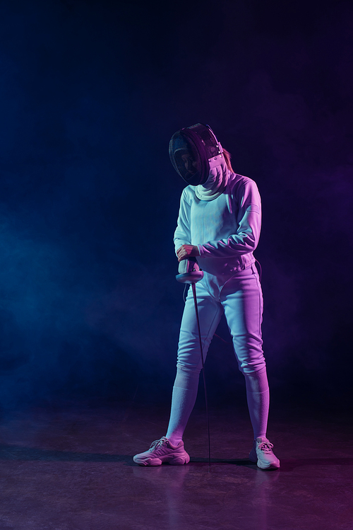 Fencer in fencing mask, suit and glove holding rapier on black background with smoke