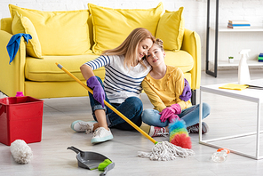 Tired mother hugging daughter with cleaning supplies on floor near coffee table in living room
