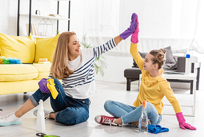 Mother and daughter with cleaning supplies giving high five, smiling and looking at each other on floor in living room