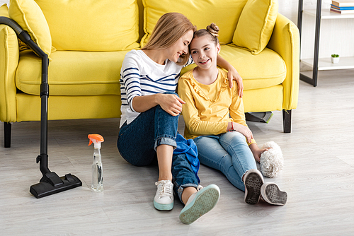 Mother hugging cute daughter and smiling near sofa and cleaning supplies on floor in living room