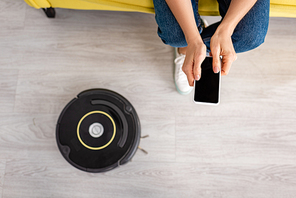 Cropped view of woman holding smartphone near robotic vacuum cleaner on floor in living room