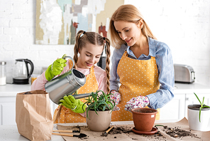 Cute child watering aloe with mother near table with paper bag, gardening tools and flowerpots in kitchen