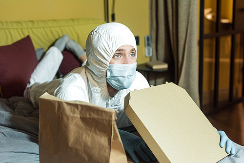 Selective focus of man in medical mask and hazmat suit watching tv on bed near package and pizza box