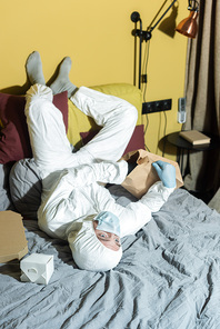 Man in medical mask, latex gloves and hazmat suit lying near packages and pizza box on bed