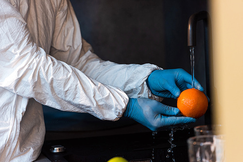 Cropped view of man in latex gloves and hazmat suit washing orange in kitchen