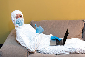Side view of man in medical mask and hazmat suit showing like sign at camera while holding laptop on couch
