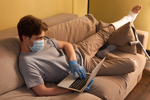 Selective focus of man in medical mask and plaster bandage on leg using laptop on couch