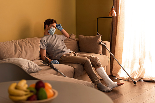 Selective focus of man in medical mask and plaster bandage on leg sitting on couch at home