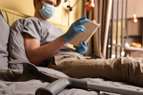 Selective focus of crutch near man in medical mask and latex gloves reading book on bed