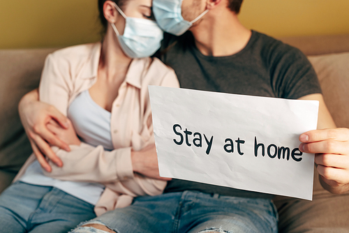 selective focus of man hugging girl in medical mask and holding placard with stay home lettering
